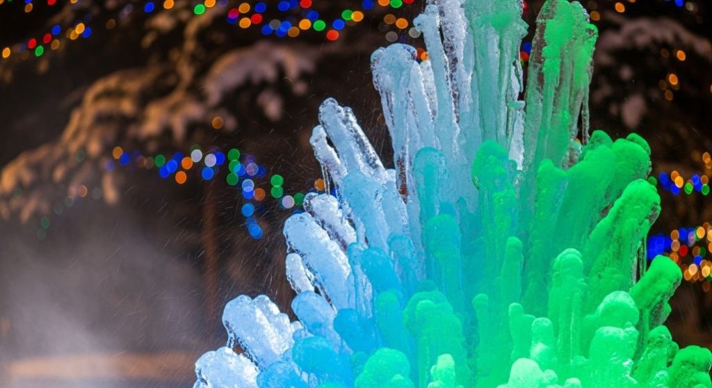 밤하늘 아래 푸른색과 초록색 조명으로 환상적으로 빛나는 거대한 얼음분수. 섬세한 얼음 결정들이 신비로운 분위기를 자아낸다.